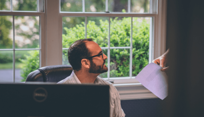A man at a computer being handed paperwork.