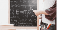 Equations on a blackboard with a teacher standing by.