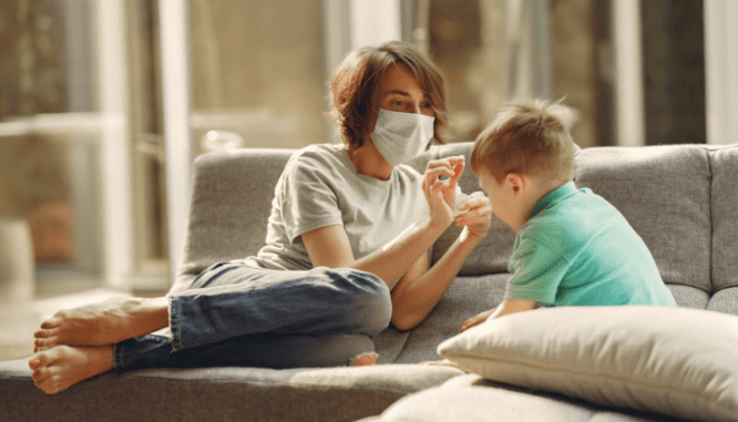 A woman sitting on a couch wearing a face mask with a toddler during coronavirus.