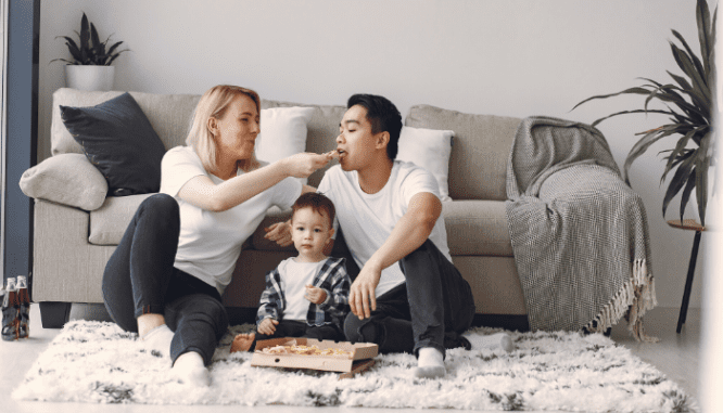 A couple with a toddler eating pizza on a shag rug in a living room.