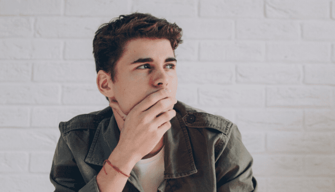 A man with brown hair and a black leather jacket holding his hand to his face while thinking about how long a house showing should take.