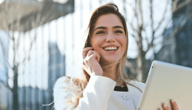 A woman smiling and listening to a phone call through a headset while holding a tablet.