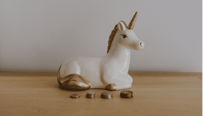 A unicorn-shaped piggy bank with stacks of coins in front of it.