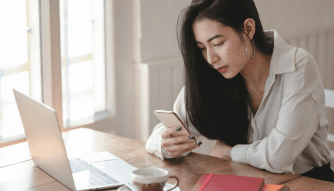 A woman looking at her phone while seated in front of a laptop.