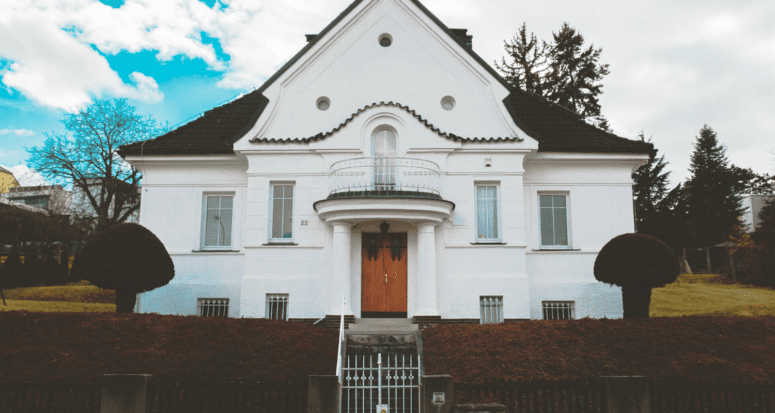 A grand white house with hedges and two stories.