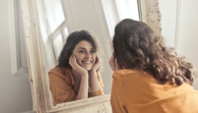 A brown haired woman wearing a yellow shirt and smiling into a mirror she can hang for home improvement in a small home.