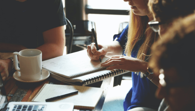 Someone in a blue shirt writing in a paper notebook with a pen on a table.