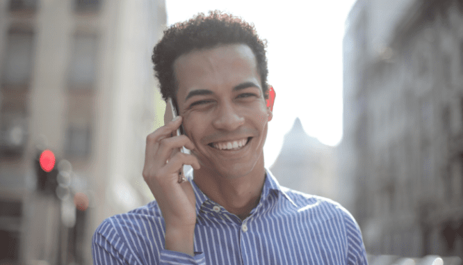 A man with brown hair and a blue striped shirt smiling while talking on the phone.