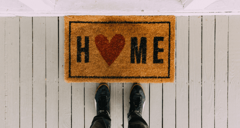 Feet standing at a welcome mat that says "Home" with a heart for the O, on a white porch