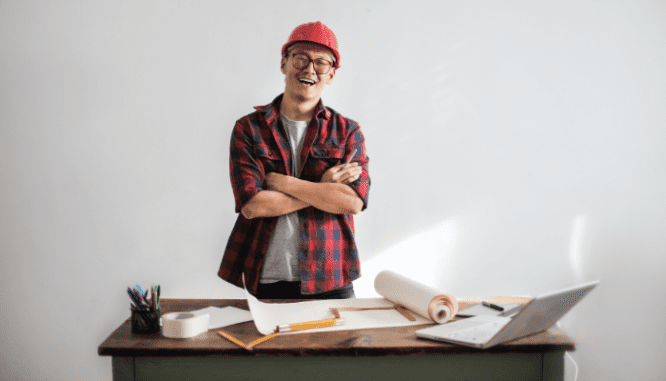 A contractor with a red helmet and a plaid shirt crossing his arms and smiling with construction plans and supplies in front of him.