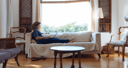 A woman sitting on a couch in a cream and wood-themed living room, using a laptop to look for a contractor.