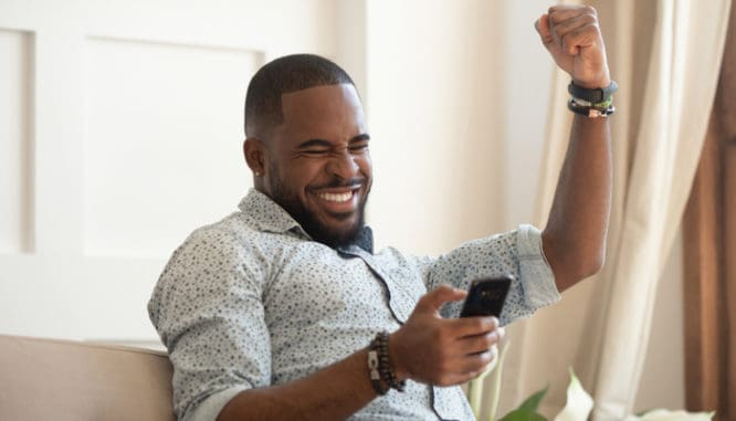 A happy Black man with a short beard looking at his phone and raising one arm in victory because his all cash offer was accepted