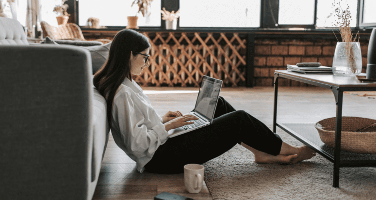 A woman sitting on the floor of a white living room, using a computer to buy a house online.