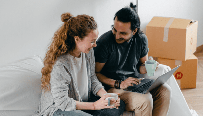 A couple using a computer to buy a house online, with several packing boxes behind them.