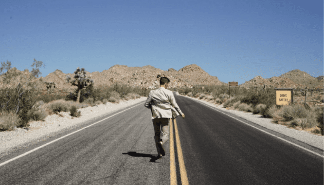 A man in a gray suit running down a desert highway, in order to go live off the grid.