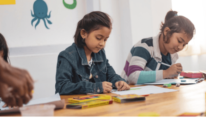 A little girl sitting at a school desk and using colored pencils to work on something in front of her.