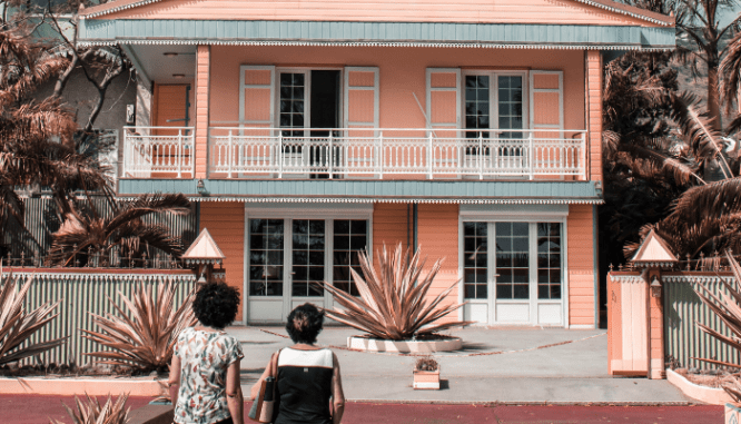 Two middle aged women standing in front of a pink house with white shutters and balconies.