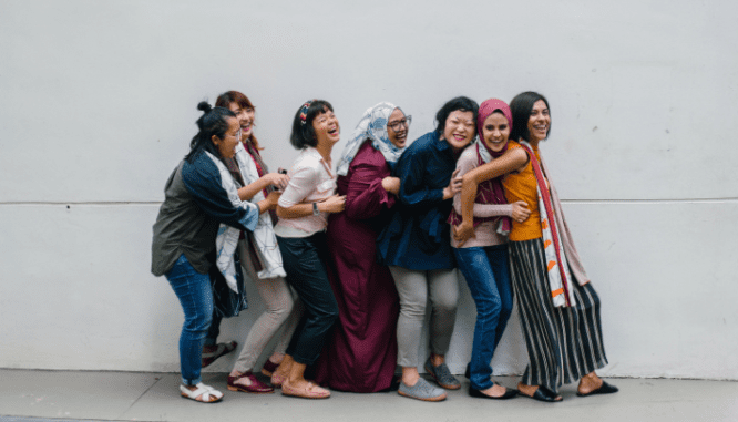 A line of women from varying ethnicities and wearing different outfits from jeans to head veils are smiling and looking forward to working against interlocking oppressions in real estate.