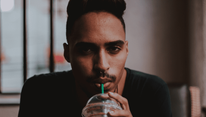 A Black or ethnic man with a septum piercing and a black sweater is staring forward, drinking from a straw.