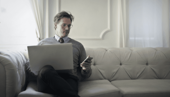 A businessman sitting on a white couch with a computer on his lap. He is looking at his smart phone, perhaps making an offer on a house.