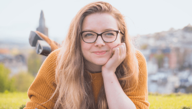 A blonde woman wearing a yellow sweater with glasses leaning on her hand and smiling at the camera after making an offer on a house.