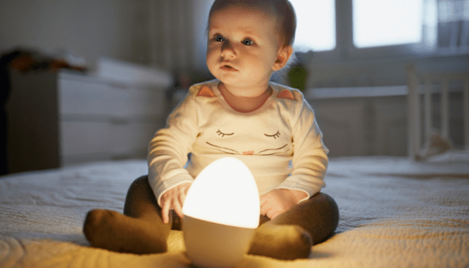 An infant wearing a kitten pajama shirt and sitting on a bed. There is a nightlight in front of them.