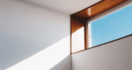 A wooden trimmed window with blue sky letting a beam of sunlight onto a white wall.