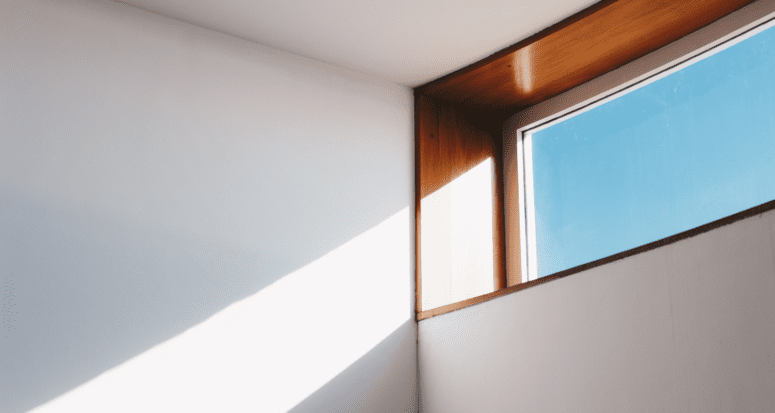 A wooden trimmed window with blue sky letting a beam of sunlight onto a white wall.