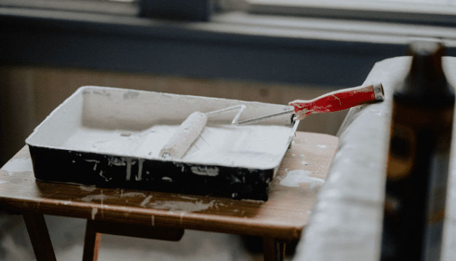 A roller brush in a pan of white interior paint being used by a homeowner working on how to increase home value for appraisal.