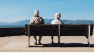 An elderly couple with dementia sitting on a bench near their house.