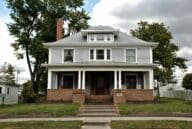 Gray American Foursquare house with a white trim and a covered porch and a brick foundation.