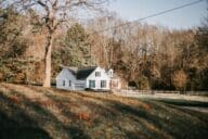 Small white farmhouse in South Carolina with black shutters and a wood barn, seen from a distance and surrounded by trees and yellowing grass.