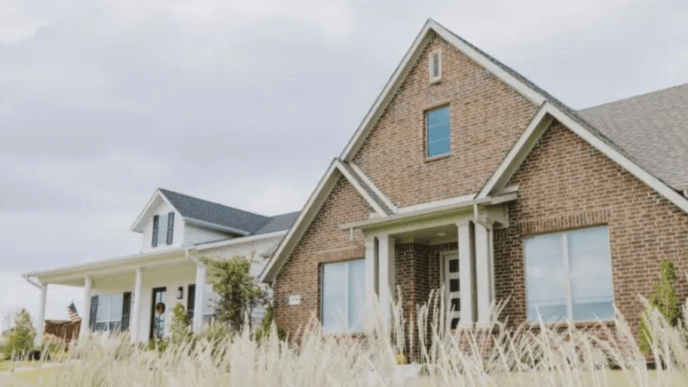 A brick house and a white house next door to each other with tall grass in the foreground.