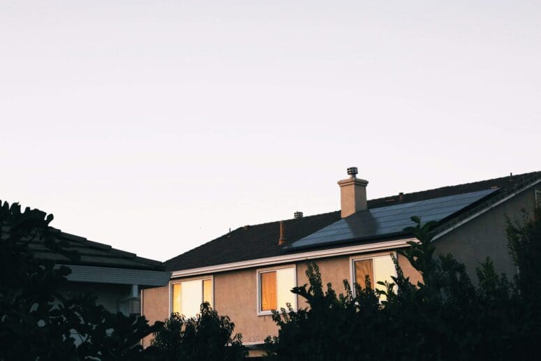 Tan stucco house with solar panel on the roof and shadowy greenery in the foreground.