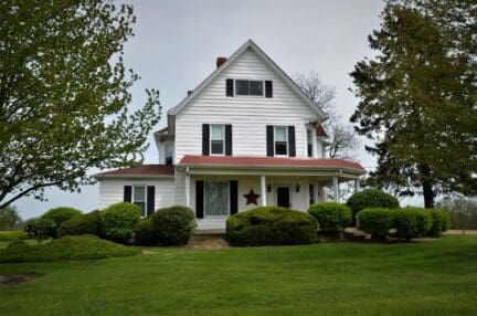 White farmhouse with a red shingle roof on a grassy lawn. There's a large metal star decoration on the first floor of the house near the front door.