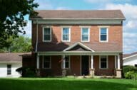 Two-story brick farmhouse with white trim, a covered front porch, and an American flag flying by the front door.