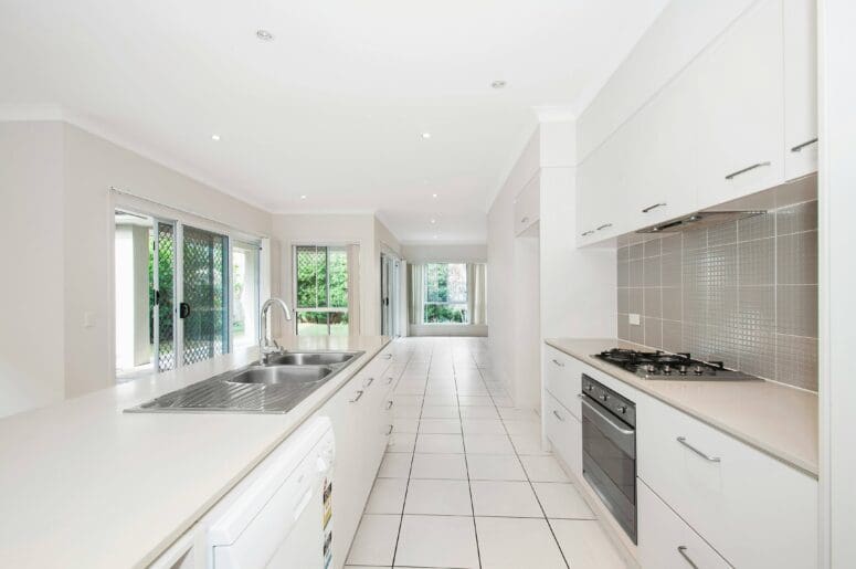 A clean, empty house with an all-white kitchen and a straight shot to the dining space, which has white square tile flooring and large windows.