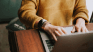 Anonymous person wearing yellow sweater and bracelets typing on a laptop on a wooden table.