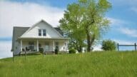 White two-story farmhouse on a grassy hill with a fire fence in the foreground. There is a white rocking chair on the covered front porch.