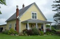 Yellow two-story farmhouse with front porch trimmed in white. The garden beds surrounding the house are lush with plants and flowers.