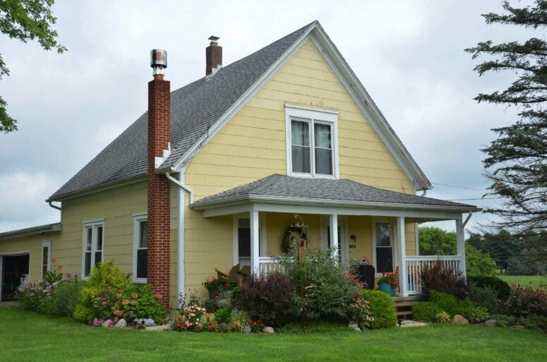 Yellow two-story farmhouse with front porch trimmed in white. The garden beds surrounding the house are lush with plants and flowers.