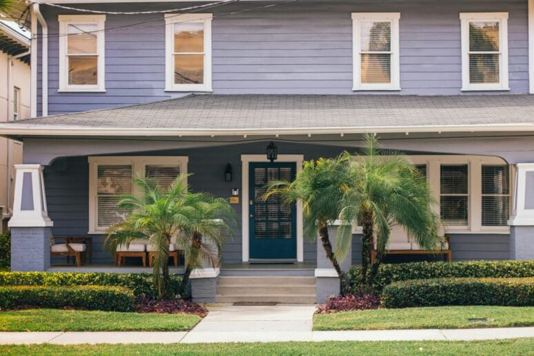 A violet craftsman-style house in Tampa with a dark blue, paned-glass front door, and small palm trees framing the entrance to the house.