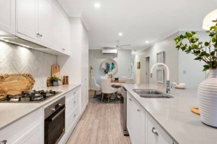 White kitchen with light gray countertops and a herringbone-patterned backsplash. The kitchen overlooks a small dining area with a round table and four upholstered chairs.