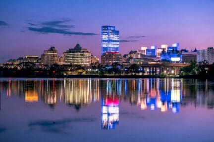 The skyline of Albany, New York at twilight with reflections of buildings and their lights seen in the water of the Hudson River.