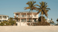 A light pink two-story beach house in Madeira Beach, Florida, surrounded by sand and towering palm trees.