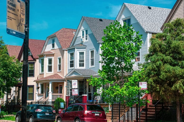 A row of houses with cars parked in front of them in Logan Square, Chicago. The houses are pastel-colored, three stories, and have covered front porches.